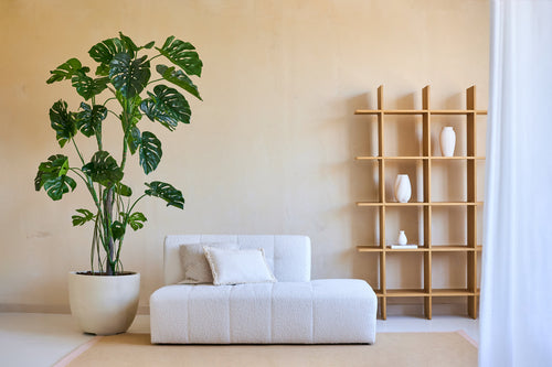 Salón minimalista con una planta Monstera artificial de 240 cm en maceta, un moderno sofá blanco con cojín y un armario abierto de madera con jarrones blancos contra una pared beige. Luz natural desde la derecha.