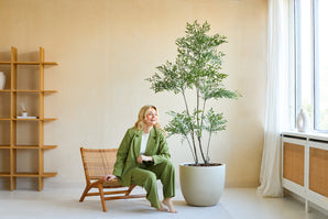 Una mujer con traje verde sonríe sentada en una silla de madera junto a la planta Ruscus Artificial de 180 cm en una habitación luminosa y moderna con paredes beige, grandes ventanales y decoración minimalista.