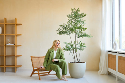Una mujer con traje verde sonríe sentada en una silla de madera junto a la planta Ruscus Artificial de 180 cm en una habitación luminosa y moderna con paredes beige, grandes ventanales y decoración minimalista.