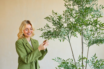 Een glimlachende vrouw in een groene blazer staat binnen en raakt voorzichtig de bladeren aan van een Kunstplant Ruscus 150 cm met dunne takken en weelderig groen gebladerte, dat afsteekt tegen een effen, lichtgekleurde achtergrond.