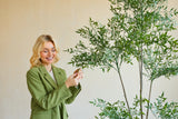 Una mujer sonriente con una chaqueta verde está en el interior y toca suavemente las hojas de una planta de Ruscus artificial de 150 cm con ramas delgadas y follaje verde exuberante, que destaca sobre un fondo liso y de color claro.