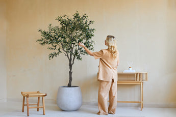 Een vrouw in losse beige kleding staat binnen naast een Kunst Olijfboom van 200 cm en raakt zachtjes de bladeren aan. Vlakbij staan een houten kruk en een consoletafel met boeken en een diffuser tegen de beige muur.