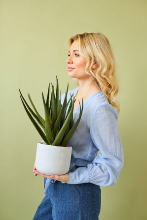 Una mujer de pelo largo y rubio, con blusa azul claro y pantalón azul, sostiene la planta artificial Aloe Vera de 43 cm en una maceta sobre un fondo liso de color verde claro.