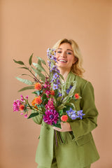 Een lachende blonde persoon in een groen pak houdt het levendige Zijden kunstbloemen boeket Colourful Svenski tegen een effen, lichtbruine achtergrond.