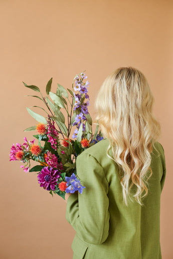 Een persoon met lang, golvend blond haar en een groene jas is van achteren gezien, terwijl hij het Zijden kunstbloemen boeket Colourful Svenski vasthoudt tegen een effen beige achtergrond.