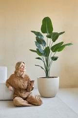 Una mujer con un mono marrón está sentada sonriendo en el suelo con una taza de café a su lado, junto a la Planta Artificial Strelitzia 180 cm real touch en una habitación minimalista con paredes beige.