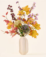 El ramo de flores artificiales de seda 'Wild Amethyst', con hojas otoñales, flores violetas y blancas y hortensias verdes, se presenta en un jarrón de cerámica blanca sobre un fondo liso y claro.