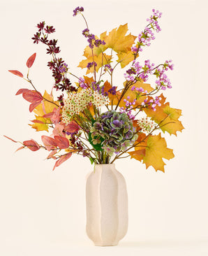 El ramo de flores artificiales de seda 'Wild Amethyst', con hojas otoñales, flores violetas y blancas y hortensias verdes, se presenta en un jarrón de cerámica blanca sobre un fondo liso y claro.