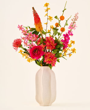 En un jarrón de cerámica blanca se encuentra el ramo de flores artificiales de seda 'Summer Blush', compuesto por flores rosas, amarillas y naranjas con hojas verdes, sobre un fondo liso y claro.