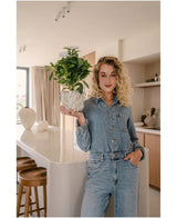 Een vrouw met krullend blond haar, gekleed in een spijkerjumpsuit, staat in een moderne keuken en glimlacht terwijl ze de Kunstplant Gardenia UV 39 cm wit vasthoudt – een kleine gardenia in een pot met witte bloemen. Op de achtergrond zijn twee barkrukken en een minimalistische inrichting te zien.