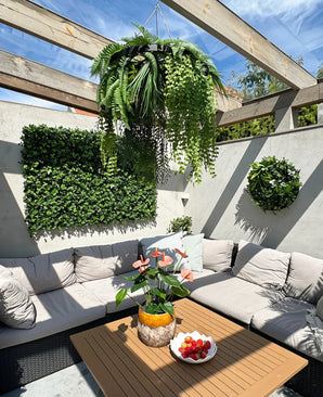 Una terraza soleada con muebles para sentarse en color beige con cojines y una mesa de madera, decorada con un jarrón de piña con flores rosas y un cuenco de fresas. Las plantas de pared verdes crean un interior exuberante y se complementan con la planta colgante artificial helecho UV de 60 cm en anillo negro como llamativo acento colgante.