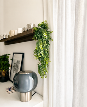 Un salón moderno con una lámpara de cristal gris, cuadros enmarcados, una maceta y un estante de madera con tazas y la planta colgante artificial 'Pannenkoek' (61 cm), cuyos zarcillos cuelgan junto a cortinas blancas.