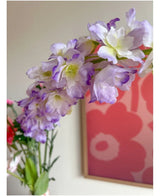 Primer plano de la flor artificial Delphinium violeta de 105 cm, dispuesta en un jarrón, con una obra de arte floral rosa y rojo en la pared del fondo.