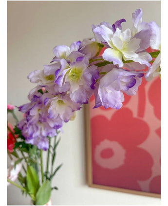Primer plano de la flor artificial Delphinium violeta de 105 cm, dispuesta en un jarrón, con una obra de arte floral rosa y rojo en la pared del fondo.