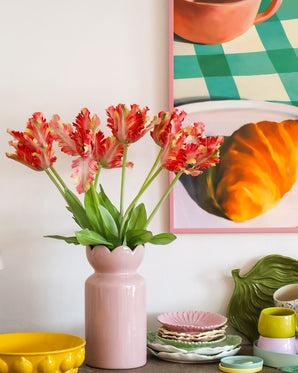 Sobre una mesa con vajillas coloridas se encuentra un jarrón rosa que contiene la flor artificial Tulipán periquito francés de 76 cm rojo/amarillo, mientras que al fondo se ve parcialmente un cuadro enmarcado de un croissant y una taza.