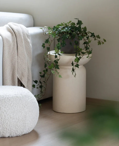 Una hiedra artificial Deluxe Green de 90 cm se encuentra en un jarrón beige sobre un suelo de madera junto a un sofá gris claro con cuadros color crema y un puf estructurado blanco.