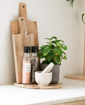 Sobre una encimera blanca, una bandeja redonda de madera contiene dos tablas de cortar de madera, dos molinillos de especias, un Art Pumila en una olla UV de 25 cm y un mortero de mármol blanco.