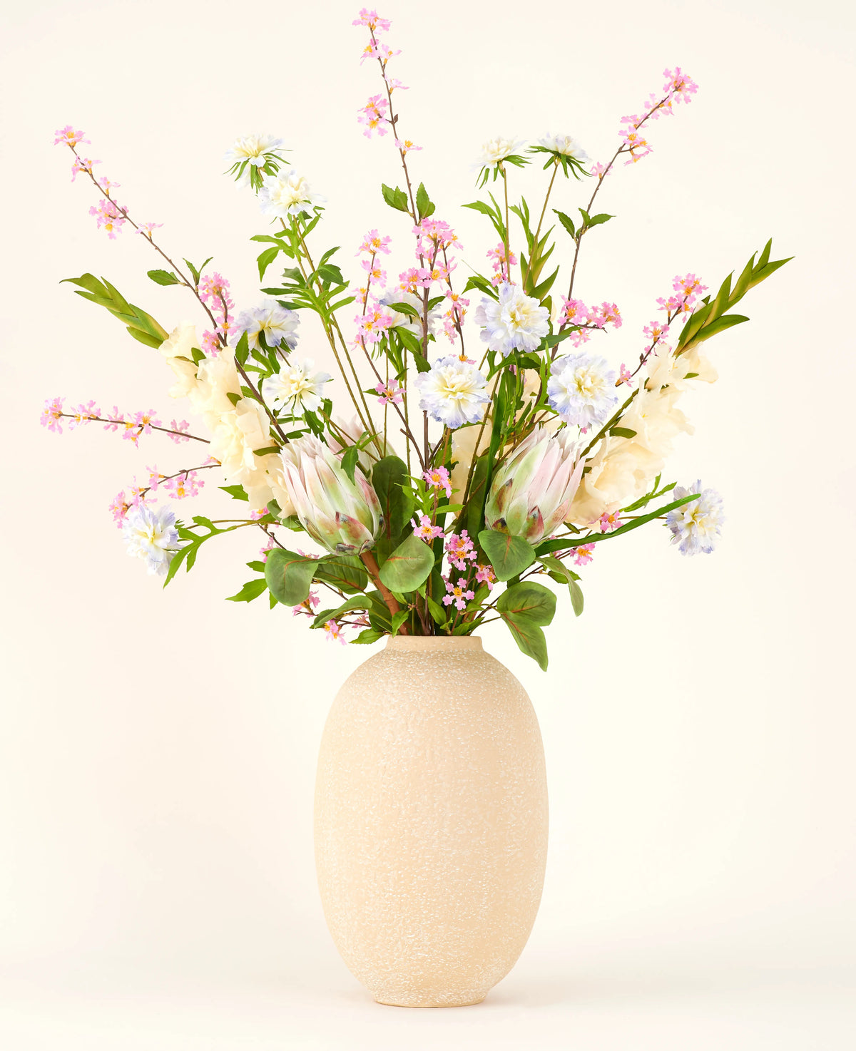 En un jarrón con estructura beige se encuentra el ramo de flores artificiales de Seda Lots of Love XL, un ramo atado a mano de flores de seda rosas, blancas y azules con hojas verdes y tallos largos sobre un fondo claro.