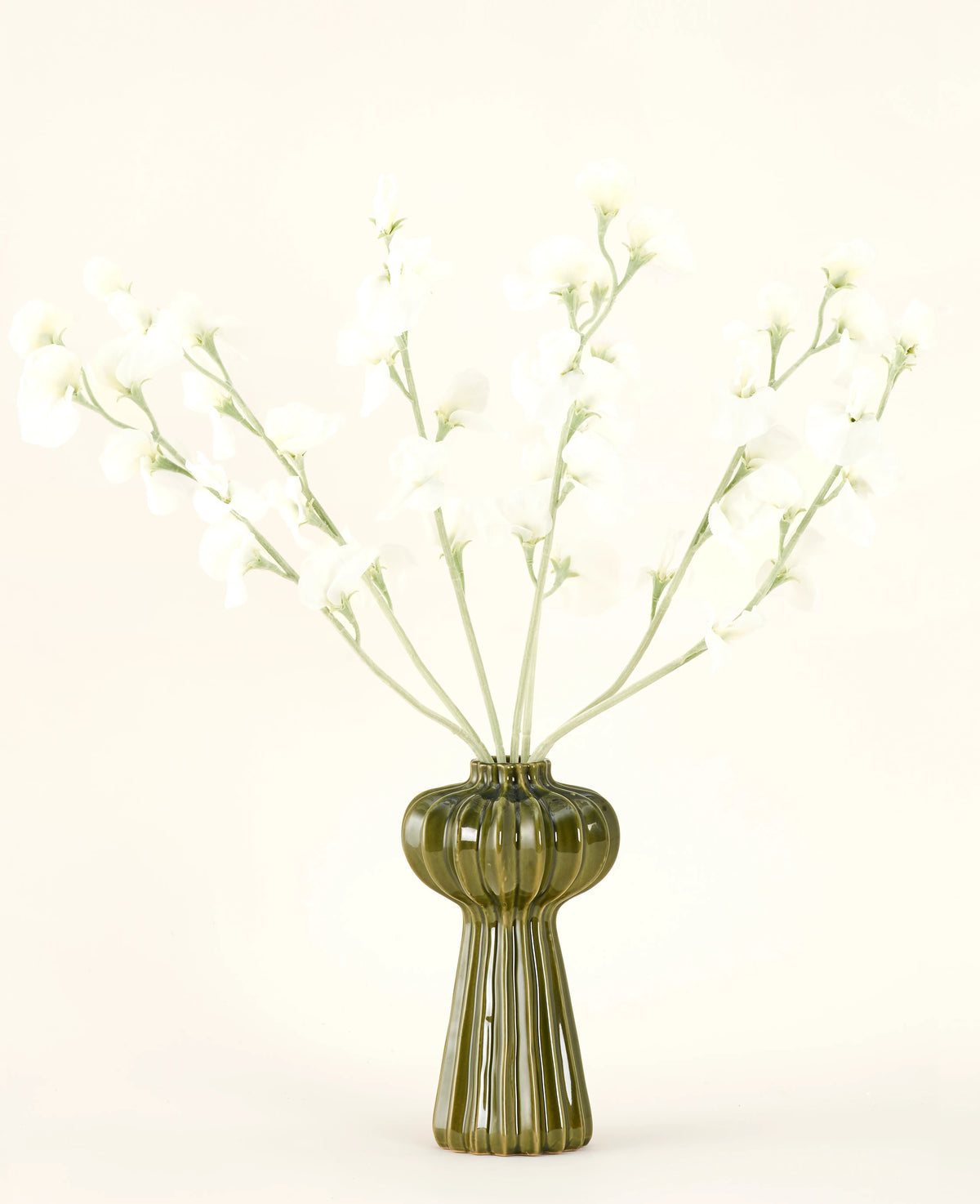 Un jarrón de vidrio acanalado verde sostiene el ramo de flores artificiales de seda color crema guisante con delicadas flores de seda blanca y tallos delgados, sobre un fondo liso blanquecino.