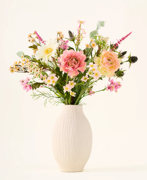 En un jarrón de cerámica blanca se encuentra el ramo de flores artificiales de seda 'Backyard Blossom', que consiste en un hermoso arreglo de rosas rosadas, margaritas blancas y pequeñas flores amarillas, hecho de flores de seda, complementado con exuberantes hojas verdes sobre un fondo liso y claro.
