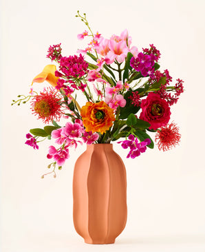 En un jarrón de color melocotón se encuentra el ramo "Tropical Sunset" de flores artificiales de seda, compuesto por una alegre mezcla de flores rosas, naranjas, amarillas y rojas sobre un fondo liso y claro.