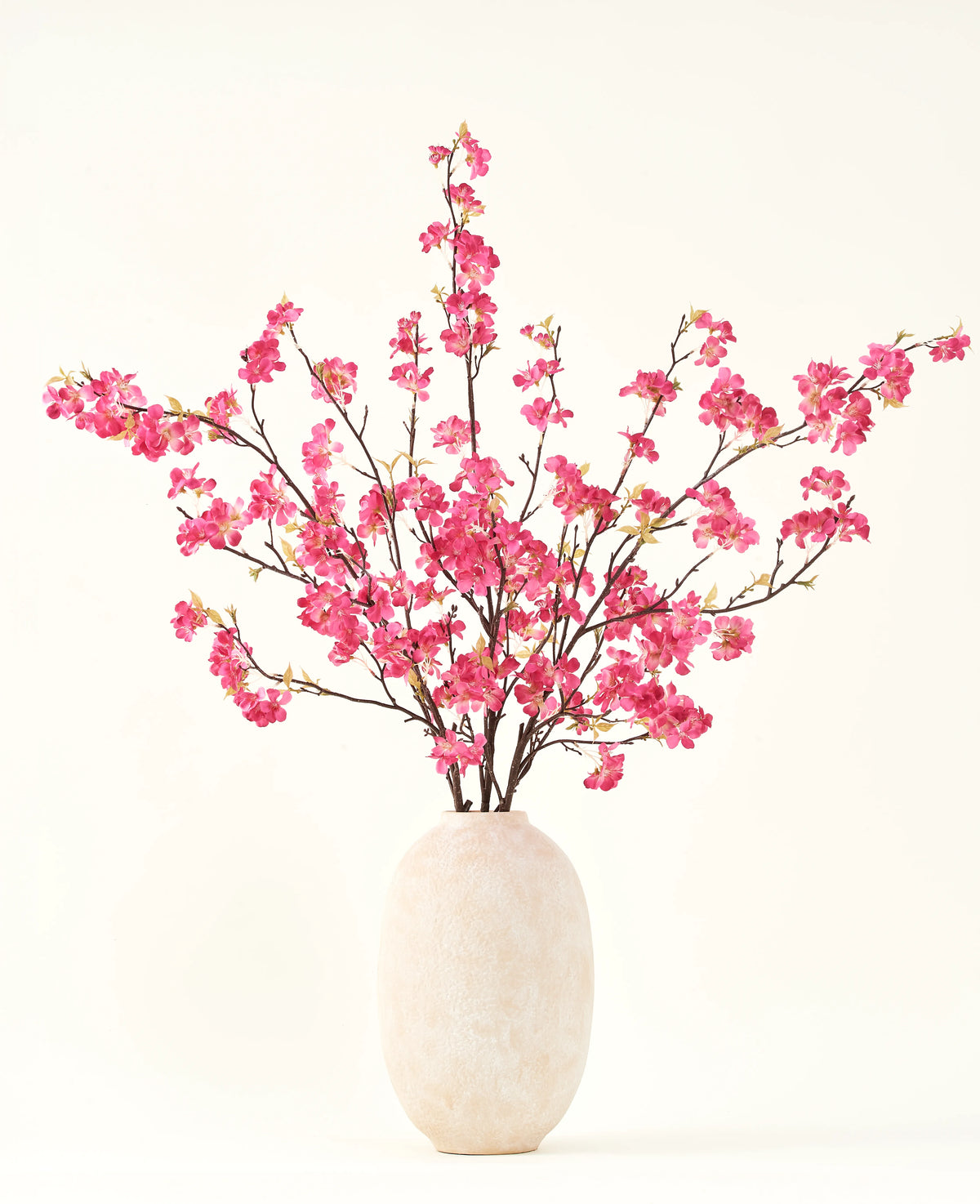 El ramo de flores artificiales de seda Flor de cerezo fucsia de 150 cm presenta vibrantes tallos de flores de cerezo fucsia con hojas verdes en un sencillo jarrón blanco, que complementará cualquier interior sobre un fondo claro.
