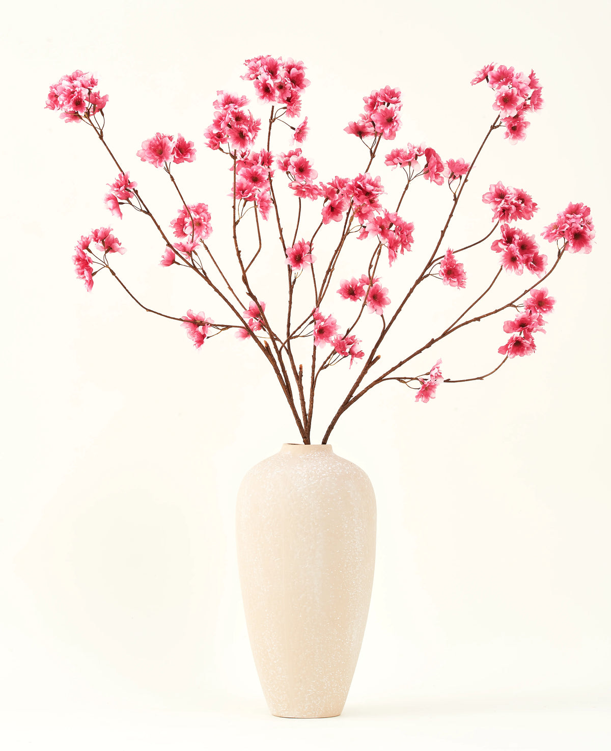 El ramo de flores artificiales de seda Flor de cerezo fucsia de 114 cm añade un vibrante toque de color a tu interior con sus llamativas ramas y flores fucsias, bellamente expuestas en un jarrón de cerámica beige sobre un fondo liso blanquecino.