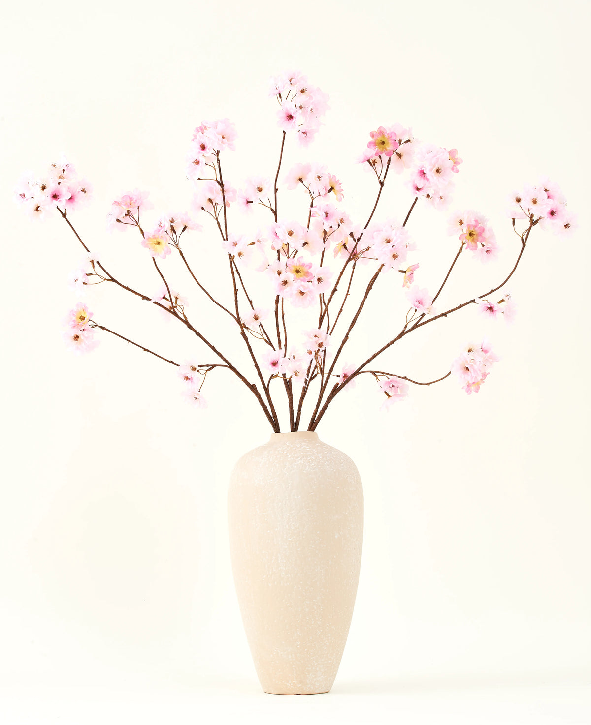El ramo de flores artificiales de seda Flor de cerezo rosa 114 cm tiene ramas largas y delgadas con racimos de flores de color rosa claro en un jarrón de cerámica beige, que forman un llamativo contraste con el fondo blanco liso.