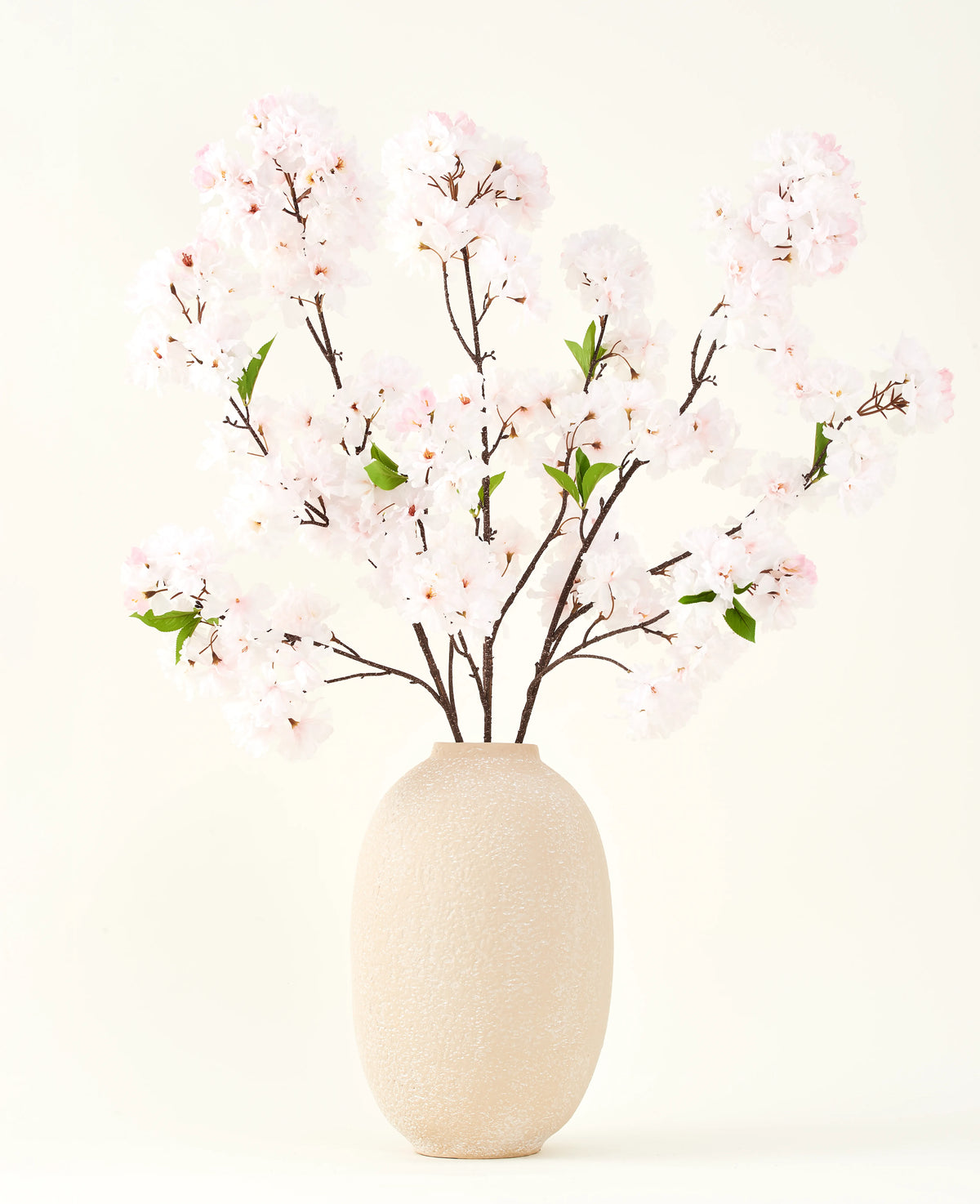El ramo de flores artificiales de seda Cherry Blossom Blush 119 cm muestra delicadas flores de cerezo de color rosa claro con hojas verdes en un jarrón beige, ideal como decoración floral artificial sobre un fondo claro.