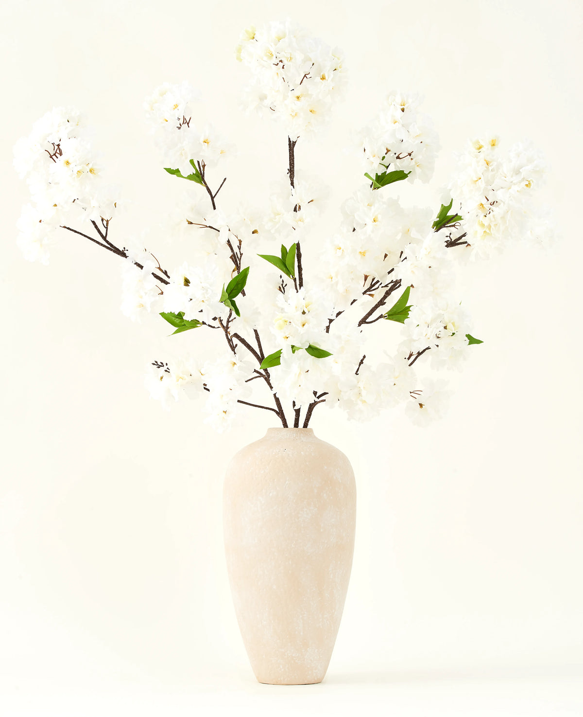 El ramo de flores artificiales de seda Crema de cerezo de 119 cm tiene elegantes ramas marrones con suaves flores artificiales blancas y hojas verdes, expuestas en un sencillo jarrón beige sobre un fondo liso y claro.
