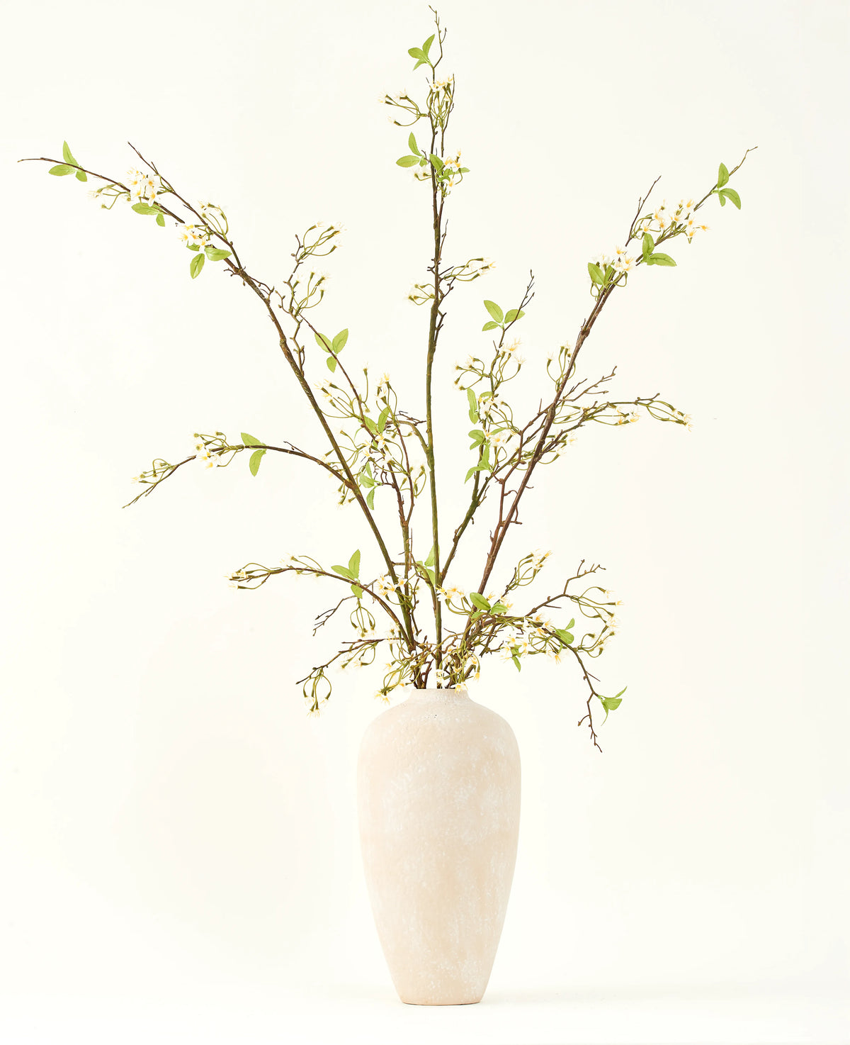 Un jarrón alto de cerámica de color crema contiene el ramo de flores artificiales de seda Verbena crema con largas ramas, pequeñas hojas verdes y delicadas flores blancas, bellamente dispuestas sobre un fondo liso y claro.