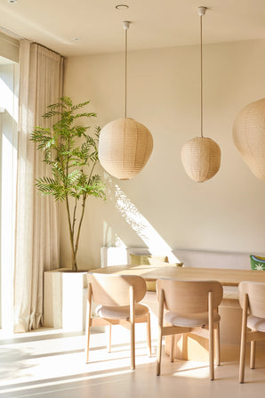 Un comedor luminoso y minimalista está amueblado con sillas de madera clara, una mesa, tres lámparas colgantes de papel, la planta Mahonia artificial de 210 cm en una maceta y cortinas de piso a techo cerca de una gran ventana que deja entrar la luz del sol.