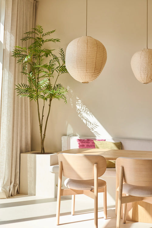 Un salón luminoso y minimalista con sillas de madera, un sofá blanco con cojines de colores, amplios ventanales con cortinas, dos lámparas colgantes farolillos de papel redondos y la Planta Artificial Mahonia de 210 cm, que aporta un toque de verde. La luz del sol proyecta sombras en las paredes.