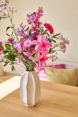 En un jarrón de cerámica blanca se encuentra el ramo de flores artificiales de seda 'Purple Dawn', un alegre arreglo de flores rosas, violetas y magenta; el jarrón se encuentra sobre una mesa de madera clara con cojines de colores al fondo.