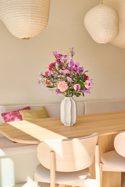 Sobre una mesa de comedor de madera clara hay un jarrón blanco con el ramo de flores artificiales de seda Purple Dawn, repleto de vibrantes tonos rosados ​​y morados. Detrás, un banco tapizado con cojines y dos faroles de papel colgantes completan la escena, impregnando el espacio de la atmósfera tranquila inspirada en Purple Dawn.