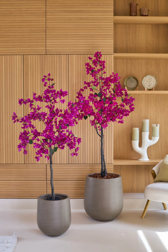Twee potplanten van het type Kunstplant Bougainvillea van 180 cm, in de kleur paars, met levendige paarse bloemen staan voor een lichte houten wand en planken met decoratieve voorwerpen zoals kaarsen en borden. Rechts staat een beige stoel, die een gezellige sfeer toevoegt aan dit levendige kunstplant-woonkamertje.