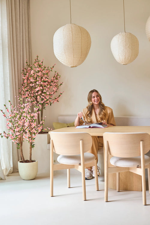 Una mujer está sentada sonriendo ante una mesa con un libro abierto. La habitación está decorada con muebles de madera clara, faroles de papel, una suave luz natural que entra a través de cortinas altas y una planta de cerezo artificial rosa de 120 cm que añade un toque de flor rosa.