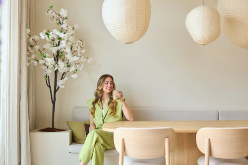 Una mujer con un traje verde claro está sentada en un banco junto a una mesa de comedor de madera, con una taza en la mano. Sobre ella cuelgan farolillos de papel decorativos y en la esquina se alza la flor de cerezo artificial blanca de 180 cm, mientras que una suave luz natural entra a raudales a través de una ventana.