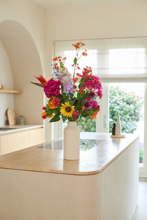 Un jarrón blanco con el ramo de flores artificiales de seda 'Colourful Spirit' se encuentra sobre una isla de cocina de madera clara en una cocina moderna y luminosa con grandes ventanales y una decoración neutra, creando un punto focal sustentable y animado.