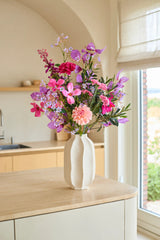 En una isla de cocina de madera clara se encuentra un jarrón blanco con el ramo de flores artificiales de seda 'Purple Dawn', compuesto por flores artificiales de color rosa y violeta, con una ventana y, al fondo, armarios de cocina beige.
