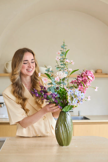 Een vrouw met lang, golvend haar schikt een boeket ‘Lavender Dreams’ van Zijden kunstbloemen in een groene vaas in haar moderne keuken, terwijl ze glimlachend aan het werk is. Het levendige boeket van zijden bloemen brengt tijdloze schoonheid in haar huis.