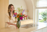 Een glimlachende vrouw die het boeket ‘Zijden kunstbloemen Colourful Svenski’ in een groene vaas schikt op een lichtgekleurd aanrecht in een keuken met een moderne inrichting en natuurlijk licht.