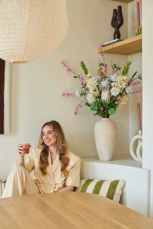 Una mujer con un traje color crema está sentada en un banco con una taza en la mano y una sonrisa en el rostro. Detrás de ella se encuentra un gran jarrón con el ramo de flores artificiales de seda 'Lots of Love XL', compuesto por flores rosas, blancas y amarillas, y al fondo estantes en los que se exponen objetos decorativos. El entorno parece acogedor y moderno.
