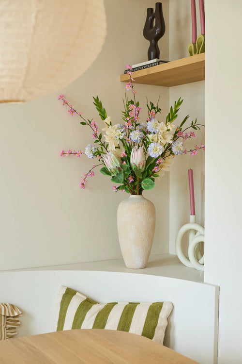 Sobre un estante blanco hay un jarrón beige con el colorido ramo de flores artificiales de seda 'Lots of Love XL', junto a velas rosas y objetos decorativos. Hay un cojín a rayas verdes y blancas sobre una silla debajo del estante.