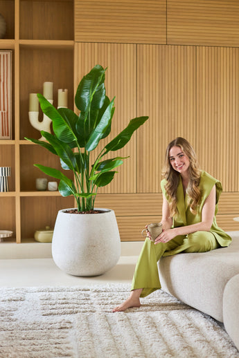 Een vrouw in een lichtgroene outfit zit op blote voeten op een bank, glimlachend en met een mok in haar hand. Naast haar staat de Kunstplant Strelitzia Real Touch Deluxe 150 cm, terwijl houten planken en decoratieve voorwerpen de achtergrond een warme uitstraling geven.