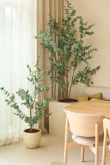 Dos plantas artificiales de eucalipto de 150 cm, colocadas junto a cortinas transparentes y paredes beige, combinadas con una mesa de comedor y sillas de madera clara en un interior minimalista y moderno.