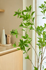 Una planta artificial Pieris Japonica de 150 cm se encuentra junto a una ventana junto a un mueble de madera clara que contiene botellas de cerámica y un molinillo de pimienta, añadiendo un toque minimalista a la habitación amplia y luminosa.