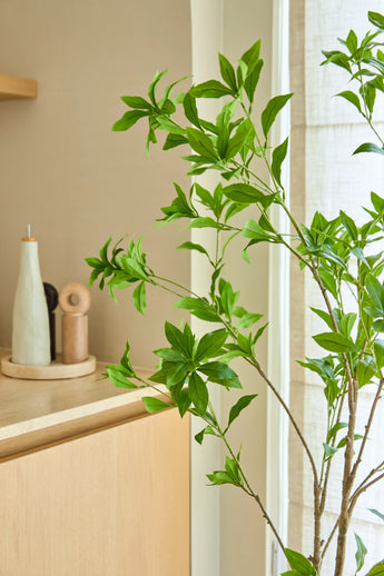 Una planta artificial Pieris Japonica de 150 cm se encuentra junto a una ventana junto a un mueble de madera clara que contiene botellas de cerámica y un molinillo de pimienta, añadiendo un toque minimalista a la habitación amplia y luminosa.