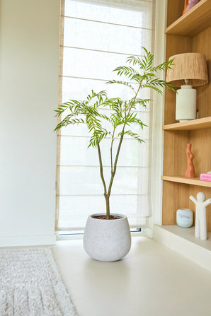 Una planta Mahonia artificial de 150 cm con esbeltas hojas verdes se alza sobre un suelo claro cerca de una ventana con persianas transparentes, junto a una estantería de madera con elementos decorativos y una suave alfombra.