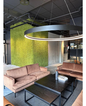 Una moderna zona de estar con sofás beige, mesas de centro negras, una gran lámpara de techo redonda y una pared verde al fondo, creada con Kunsthaag Nordic Moss 50x50 cm UV, que irradia un interior lujoso y un diseño industrial contemporáneo.